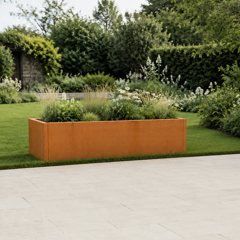A light beige stone patio in the foreground of a landscaped garden with a rust-orange rectangular planter box, green grass, shrubs, and trees. Corten steel planter tub