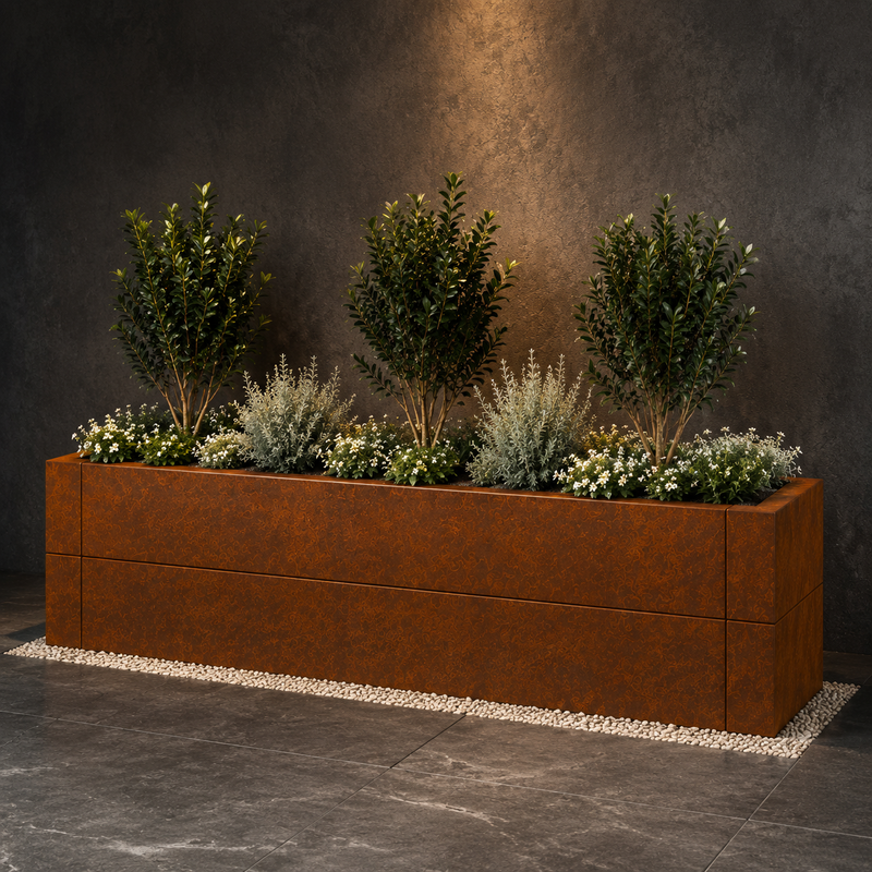 A large rectangular rusted metal planter box filled with green shrubs and small white flowers, placed on a dark stone floor against a textured dark gray wall.