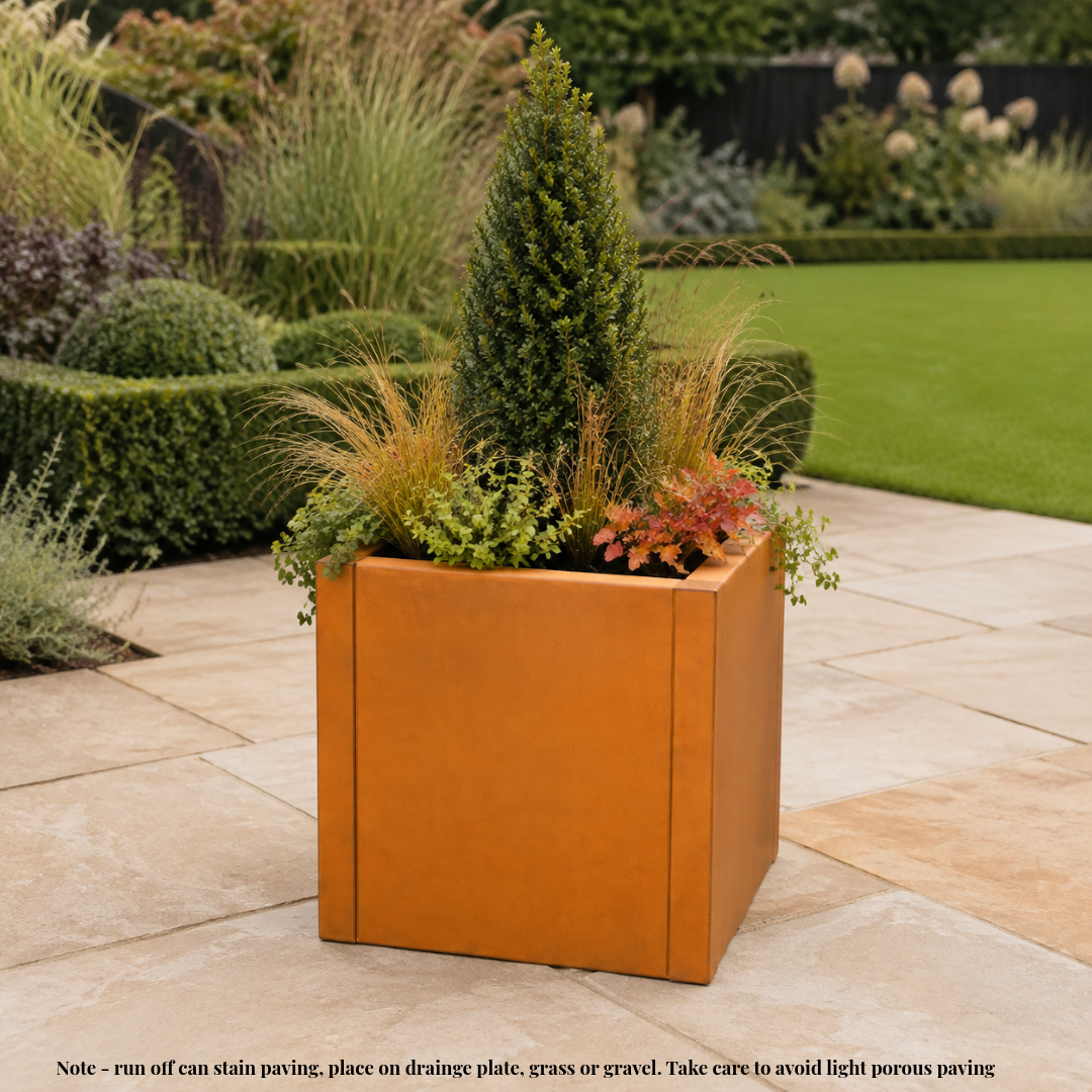 A square terracotta-colored metal planter filled with green and ornamental grass plants, placed on a light stone patio in a landscaped garden. A small note at the bottom provides care and placement  information. Rusted corten steel planter tub.