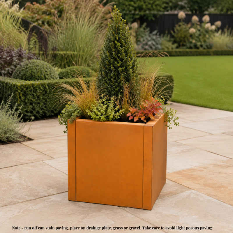 A square terracotta-colored metal planter filled with green and ornamental grass plants, placed on a light stone patio in a landscaped garden. A small note at the bottom provides care and placement  information. Rusted corten steel planter tub.