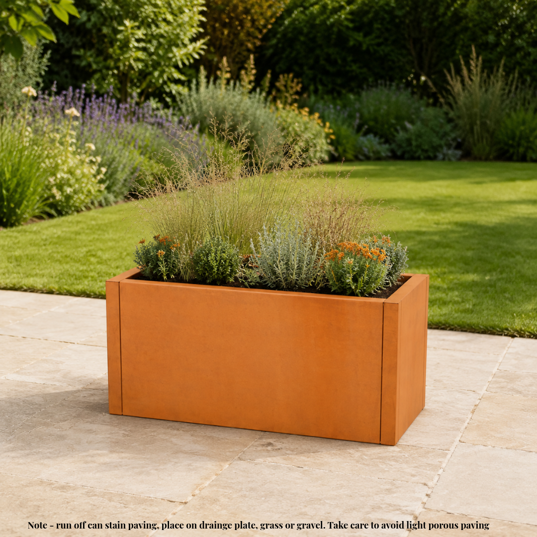 A rectangular rust-orange metal planter box filled with ornamental grasses and small plants, placed on a stone patio in a landscaped garden with green grass and shrubs in the background. Corten weathering steel