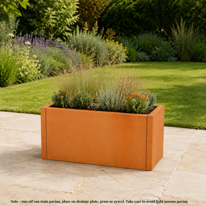 A rectangular rust-orange metal planter box filled with ornamental grasses and small plants, placed on a stone patio in a landscaped garden with green grass and shrubs in the background. Corten weathering steel