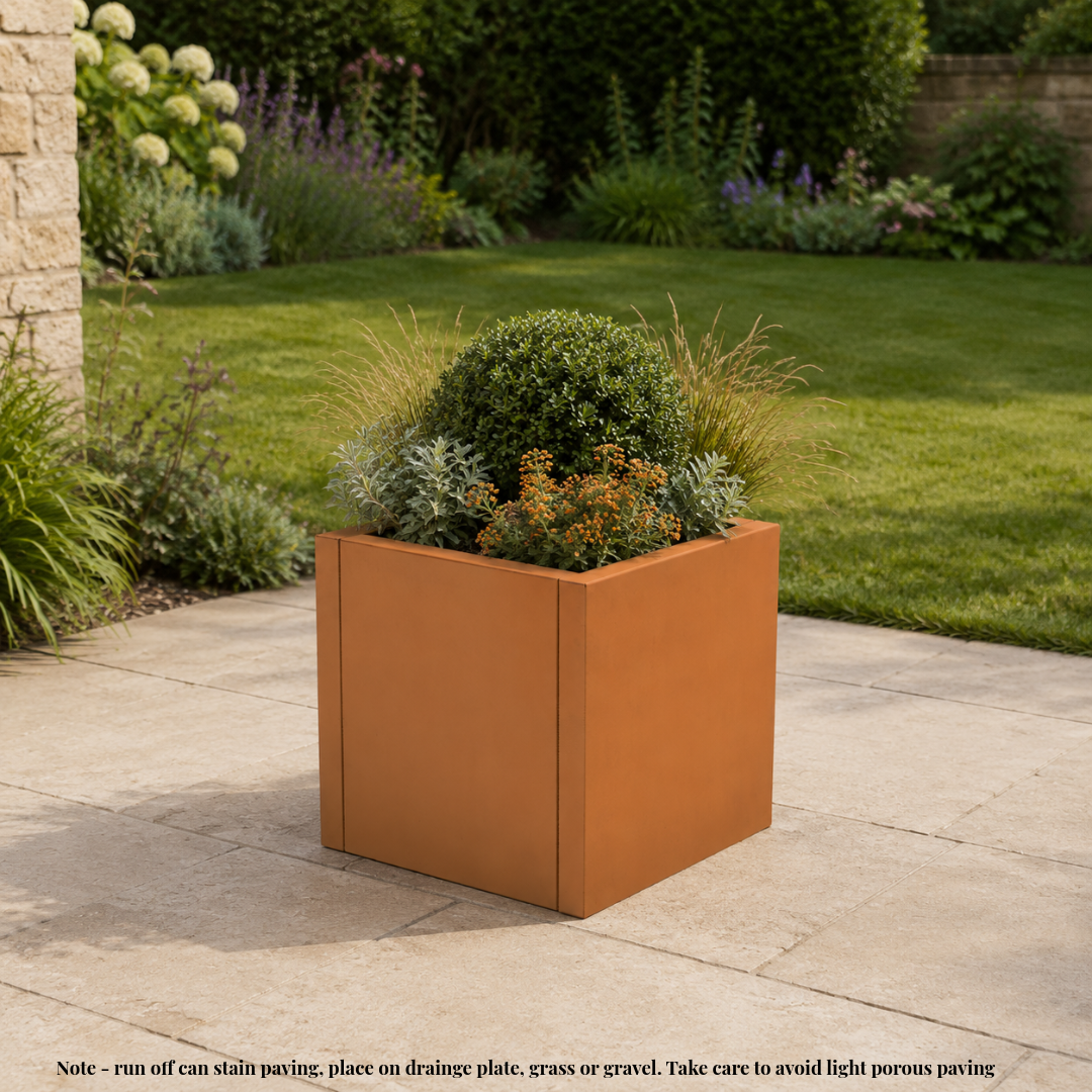 A square terracotta-colored planter box filled with green shrubs and ornamental grasses, placed on a stone patio in a landscaped garden. A small note at the bottom explains stain and drainage considerations. Corten weathering steel planter tub